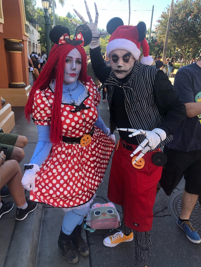 Two adults wearing costumes for Oogie Boogie Bash. They are a crossover between Mickey Mouse and Minnie Mouse and Jack and Sally from The Nightmare before Christmas.